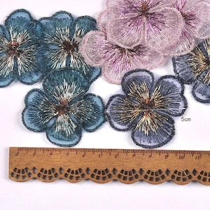 Pode incluir: Uma coleção de apliques de flores bordadas em tons de azul-petróleo e roxo claro, com detalhes em fio metálico dourado e prateado. Uma régua de madeira está posicionada abaixo das flores, mostrando que cada uma mede 5 cm de diâmetro.
