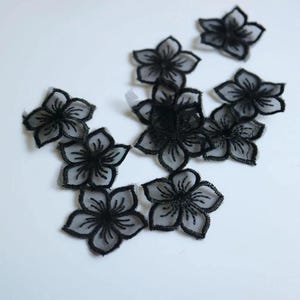 以下が含まれることがあります： 10個の黒い花形のアップリケのセット。各ピースは、半透明の透け感のある中心部と、詳細な黒い刺繍の縁取り、そして放射状に広がる花びらのラインが特徴で、繊細で重なり合った花の見た目を表現しています。