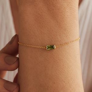 May include: A delicate gold chain bracelet featuring a single rectangular, baguette-cut olive green gemstone set in a gold bezel. The bracelet is thin and minimalist, showcasing a clear, faceted green stone centered on a fine, polished gold link chain.
