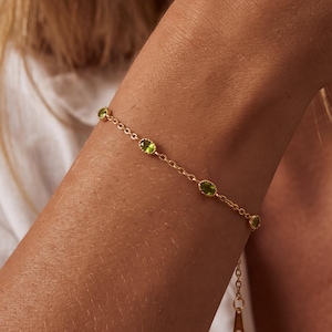 May include: A delicate gold chain bracelet featuring four evenly spaced, oval-shaped, light green peridot gemstones set in scalloped gold bezels. The bracelet includes a thin gold extender chain with a small, flat, teardrop-shaped gold charm.