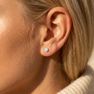 May include: A small, round, iridescent opal stud earring is centered in the earlobe. The gemstone displays a milky white base with subtle flashes of blue and green light, reflecting a soft glow against the skin of the ear.