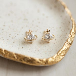 May include: A pair of gold stud earrings shaped like planets, each featuring a central round clear gemstone surrounded by a thin gold ring and topped with a small cross-shaped ornament, crafted with a polished metallic finish and clear stones.