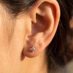 May include: A gold stud earring featuring a central round clear gemstone surrounded by a thin circular ring, resembling a planet with a small cross detail at the top. The earring is secured in the earlobe, showcasing a polished metallic finish.