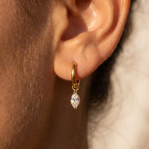 Può includere: Un orecchino a cerchio dorato con un piccolo ciondolo in pietra trasparente taglio marquise che pende dal fondo. L'orecchino è fissato al lobo dell'orecchio, mostrando una finitura metallica lucida e una pietra sfaccettata che riflette la luce.