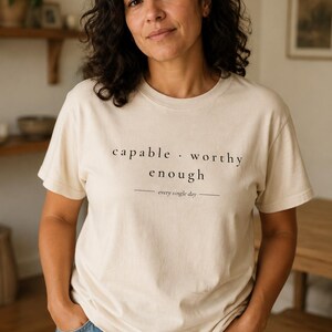 May include: A cream-colored short-sleeve t-shirt featuring black text that reads capable . worthy enough and every single day with horizontal lines on either side of the final phrase. The shirt is paired with blue denim jeans.