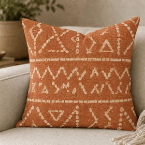 May include: A square throw pillow featuring a burnt orange base color with a distressed, off-white geometric pattern. The design consists of horizontal bands filled with repeating triangles, zig-zag lines, and dashed segments across the fabric.
