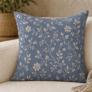 May include: A square throw pillow featuring a medium blue fabric base with a repeating off-white floral and vine pattern. The design consists of various small blossoms, leaves, and winding stems distributed evenly across the entire surface area.