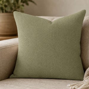 May include: A square throw pillow featuring a solid sage green color with a visible woven fabric texture. The pillow has a clean, simple design with straight edges and a uniform appearance, showcasing a consistent textile weave throughout the surface.