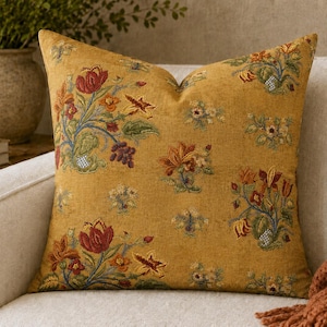 May include: A square throw pillow in a mustard yellow fabric featuring an embroidered floral pattern. The design includes various flowers and leaves in shades of red, orange, green, and blue, distributed across the front surface of the pillow.