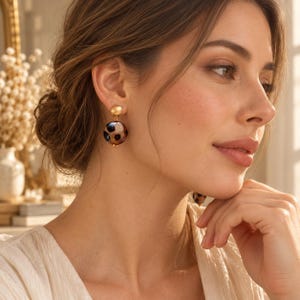 May include: A pair of drop earrings featuring a small gold-toned stud connected to a round, translucent bead with a brown and black leopard print pattern. The earrings have a polished finish and hang from the earlobe with a secure metal backing.