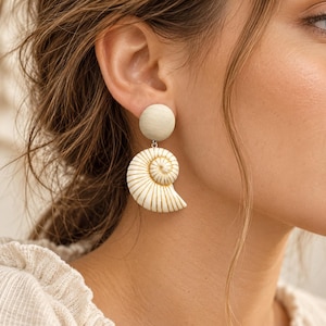 Op de afbeelding: Een hangende oorbel met een ronde, lichtbeige houten knop verbonden aan een witte nautilus schelphanger. De schelp is versierd met dunne, verticale goudkleurige lijnen die de spiraalvorm van de schelp volgen en een gedetailleerd patroon vormen.
