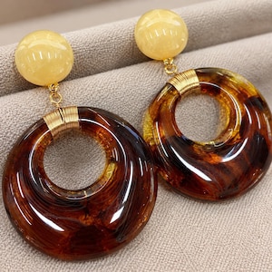 Pode incluir: Um par de brincos pendentes com um pino esférico liso de cor amarelo-pálido, ligado por um fio dourado a um grande pendente circular translúcido com padrão de tartaruga castanho e um acabamento brilhante e polido.