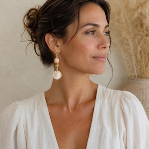 May include: A pair of drop earrings featuring a gold-tone metal post and chain. Each earring consists of a small, round, peach-colored shell bead suspended above a larger, flat, white spiral seashell with brown markings, connected by gold beads.