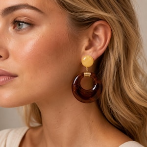 May include: A drop earring featuring a smooth, round, pale yellow stud connected by a thin gold-toned metal link to a larger, circular, dark brown translucent resin hoop with a polished finish and a hollow centre.