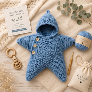 Peut inclure: Un cocon pour bébé en forme d'étoile crocheté en bleu avec une capuche et trois boutons en bois. L'article est présenté avec une fiche modèle intitulée Crochet Star Baby Cocoon, une pelote de coton bleu, un crochet en bois et un hochet.