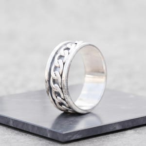 May include: A silver-toned ring featuring a central band with a raised, repeating chain-link pattern. The ring has a smooth, polished finish on the outer edges and a recessed, darker channel that highlights the intricate metal chain design.