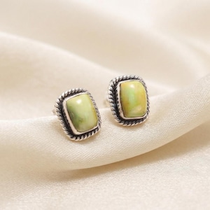 Peut inclure: Une paire de boucles d'oreilles à clous avec des pierres rectangulaires vert clair serties dans des montures en argent oxydé. Chaque pierre est entourée d'un bord en métal torsadé, créant un contour texturé qui contraste avec la surface lisse et polie des pierres cabochons vertes.