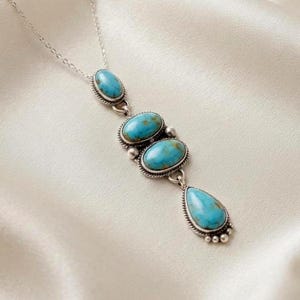 May include: A silver necklace featuring a vertical pendant with four turquoise stones. The top stone is oval, followed by two larger oval stones, and a bottom teardrop-shaped stone. Each stone is set in a silver frame with a twisted rope border.