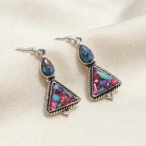 May include: A pair of silver drop earrings featuring a teardrop-shaped stone above a larger triangular stone. Both stones are inlaid with a mosaic of purple, pink, and turquoise fragments. The settings are framed with a twisted silver rope border.