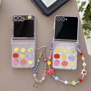 May include: Two clear foldable smartphone cases featuring colourful fruit and star stickers. One case includes a beaded wrist strap with heart and cherry charms, while the other features a longer beaded strap with star and fruit-themed cube beads.