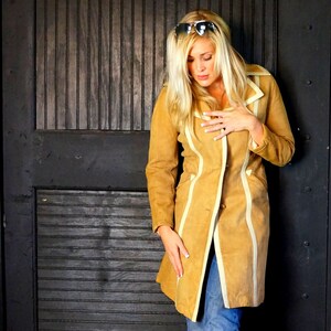 May include: A woman wearing a tan suede jacket with white trim and a pair of sunglasses. The jacket has a long, flowing style and a button closure.