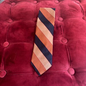 Puede incluir: Una corbata a rayas con rayas alternas azul marino, naranja y beige. La corbata está hecha de una tela suave y se muestra sobre una superficie de terciopelo rojo. Un accesorio clásico.