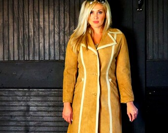 Peut inclure: Un manteau en daim beige avec des bordures blanches. Le manteau a une silhouette longue et droite et une fermeture à boutons. Le mannequin porte des lunettes de soleil et a de longs cheveux blonds.