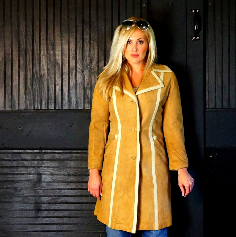 May include: A tan suede coat with white trim. The coat has a long, straight silhouette and a button closure. The model is wearing sunglasses and has long blonde hair.