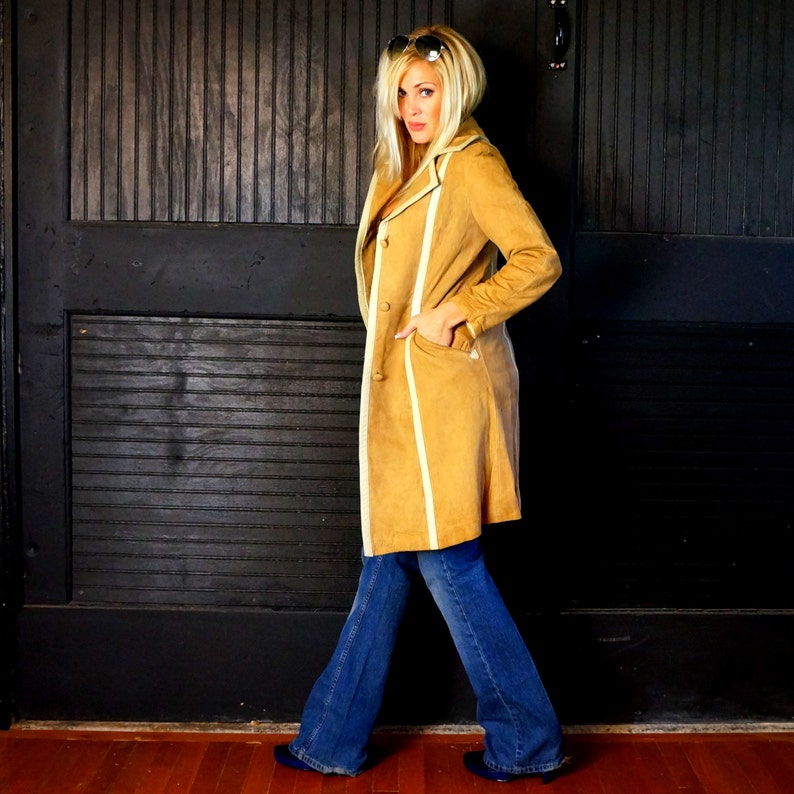 May include: A woman wearing a long, tan suede coat with white trim and blue jeans. She is standing in front of a black wooden door.