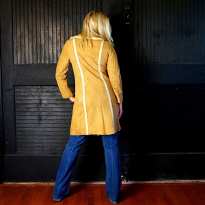 May include: A long, tan suede coat with white trim. The coat has a collar and long sleeves. The person wearing the coat is standing in front of a black door.