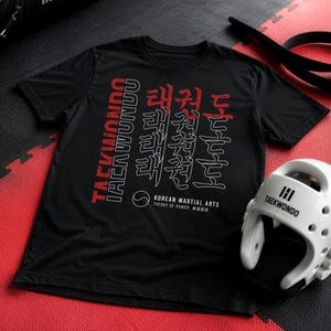 Op de afbeelding: Een zwart t-shirt met korte mouwen en een rood-witte grafische opdruk met de tekst TAEKWONDO en Koreaanse karakters. Het ontwerp bevat de tekst KOREAN MARTIAL ARTS THEORY OF POWER en een klein cirkelvormig embleem op de voorzijde.