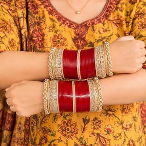 May include: A set of stacked bangles featuring deep red cylindrical bands accented by gold-toned metal spacers and intricate clear stone embellishments. The jewellery set includes multiple thin and thick bands worn together on each wrist.