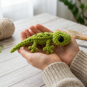May include: A small, handmade crochet lizard toy in a bright lime green colour. The lizard features a textured, knotted yarn surface, dark green spots along its back, and a single large, round, glossy black plastic eye on the side of its head.