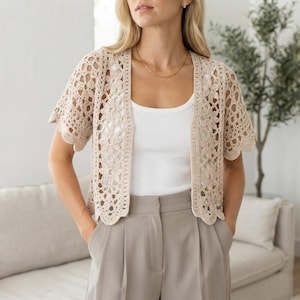 Puede incluir: Un cárdigan de ganchillo beige con un delicado patrón floral, mangas cortas y diseño frontal abierto. La prenda se lleva sobre una camiseta de tirantes blanca lisa y se combina con pantalones gris claro, mostrando un intrincado tejido calado.