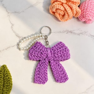 May include: A purple crocheted bow keychain featuring a silver metal ring and a short strand of white faux pearls. The bow is crafted with a textured yarn stitch and is attached to the ring by a small silver chain link.