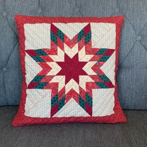 Könnte beinhalten: Quadratisches Zierkissen mit einem gesteppten Sternmuster. Der Stern besteht aus roten, cremefarbenen und grünen Blumenstoffstücken. Das Kissen hat einen roten Rand und liegt auf einer grauen Oberfläche.