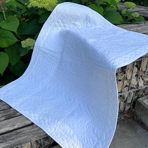May include: A white, quilted blanket with detailed floral and geometric patterns. The blanket is draped over a wooden bench, highlighting the intricate stitching and texture. The design is clean and elegant.