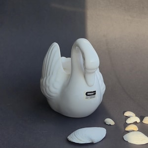 Może przedstawiać: Biała ceramiczna figurka łabędzia z formowanymi detalami skrzydeł i wygiętą szyją. Prostokątna naklejka z boku zawiera czarny napis GULDKROKEN HJO-SWEDEN. Łabędź ma wydrążony środek oraz gładkie, błyszczące wykończenie odbijające światło.