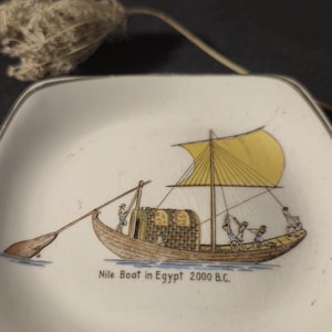Puede incluir: Un plato cuadrado de cerámica blanca con esquinas redondeadas y un borde plateado. El centro presenta una ilustración detallada de un antiguo barco del Nilo con una vela amarilla y figuras en cubierta. El texto dice: Nile Boat in Egypt 2000 B.C.