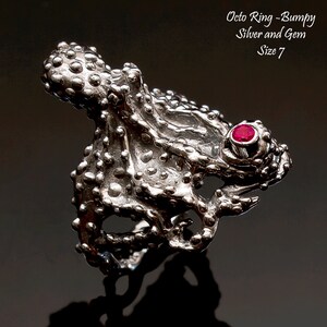 May include: A silver octopus ring with a red gem, size 7. The ring is textured and has a bumpy surface. The ring is made by Fluidbronze Studio, Sculpture and Jewelry by Rick Geib.