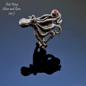 May include: A silver octopus ring with a red gem. The ring is size 7. The ring is made by Fluidbronze Studio, Sculpture and Jewellery by Rick Geib.
