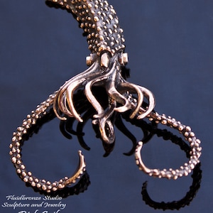 May include: Bronze giant squid pendant with ruby detailing. The pendant showcases a detailed squid design with textured skin and curling tentacles. The text "Giant Squid Pendant Bronze and Ruby" is visible.