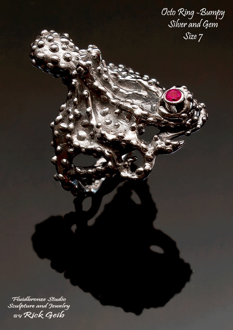 May include: A silver octopus ring with a red gem, size 7. The ring is textured and has a bumpy surface. The ring is made by Fluidbronze Studio, Sculpture and Jewelry by Rick Geib.