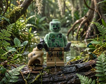 May include: A light green plastic minifigure with a printed swamp-themed torso and legs, featuring a smiling face, holding a black accessory in one hand and a grey knife in the other, positioned next to a small tan shell-like object.