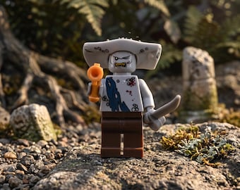 May include: A gray shark-headed minifigure with a wide, flat head, wearing a tattered white shirt with blue and red markings, and brown pants. The figure holds a small orange pistol in one hand and a curved gray sword in the other hand.