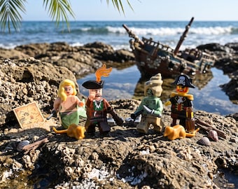 May include: Four plastic minifigures including a mermaid with blonde hair, a pirate with a flame hat, a green creature with a shell head, and a pirate with a skull hat. They stand on a rocky surface with a small treasure map and two gold rats.