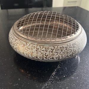 May include: A round, silver-toned metal flower frog bowl featuring an intricate, engraved floral pattern across its exterior surface. The bowl is topped with a removable, grid-patterned wire mesh dome designed for arranging and supporting flower stems.