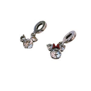 May include: Two silver-toned metal charms featuring a clear, faceted, round crystal center flanked by two smaller clear crystals. One charm includes a small, red, textured bow detail between the ears. Both charms have a circular bail at the top.