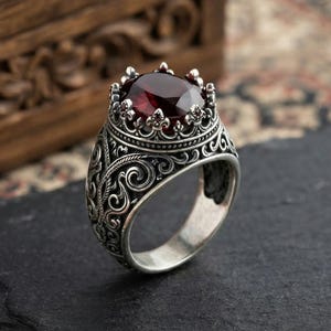 Pode incluir: Um anel de prata com um padrão de metal intrincado e em relevo ao longo da banda e uma cravação em forma de coroa. O centro do anel contém uma grande pedra preciosa vermelha escura facetada, segura por garras metálicas ornamentadas.