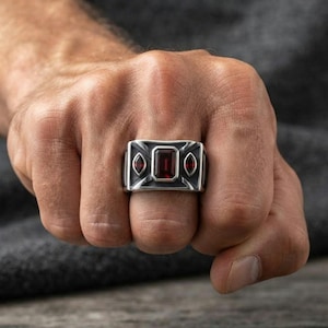 Può includere: Un anello color argento con una pietra preziosa rettangolare rosso scuro al centro, affiancata da due pietre rosse più piccole a forma di marquise. La fascia in metallo ha una finitura scura e ossidata con dettagli geometrici angolari.